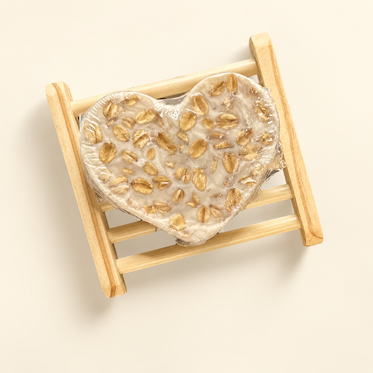 A heart-shaped soap bar wrapped in clear plastic, embedded with visible oats, displayed on a miniature wooden soap dish against a warm cream background.