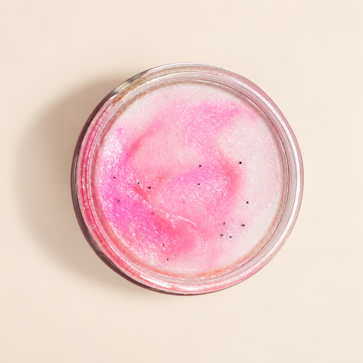 A top-down view of a clear glass jar containing a pink and white marbled sugar scrub, with tiny black specks, set against a solid beige background.