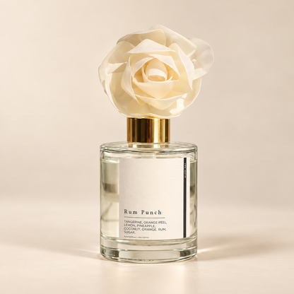 A clear glass reed diffuser bottle with a white label reading 'Rum Punch' and a large cream fabric flower on a gold cap, set against a warm beige background.