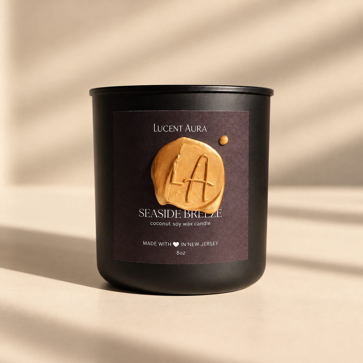 A matte black soy wax candle jar with a dark brown label reading 'Lucent Aura' and 'Seaside Breeze,' featuring a gold wax seal with an 'LA' monogram, photographed on a warm beige surface with soft diagonal shadows.