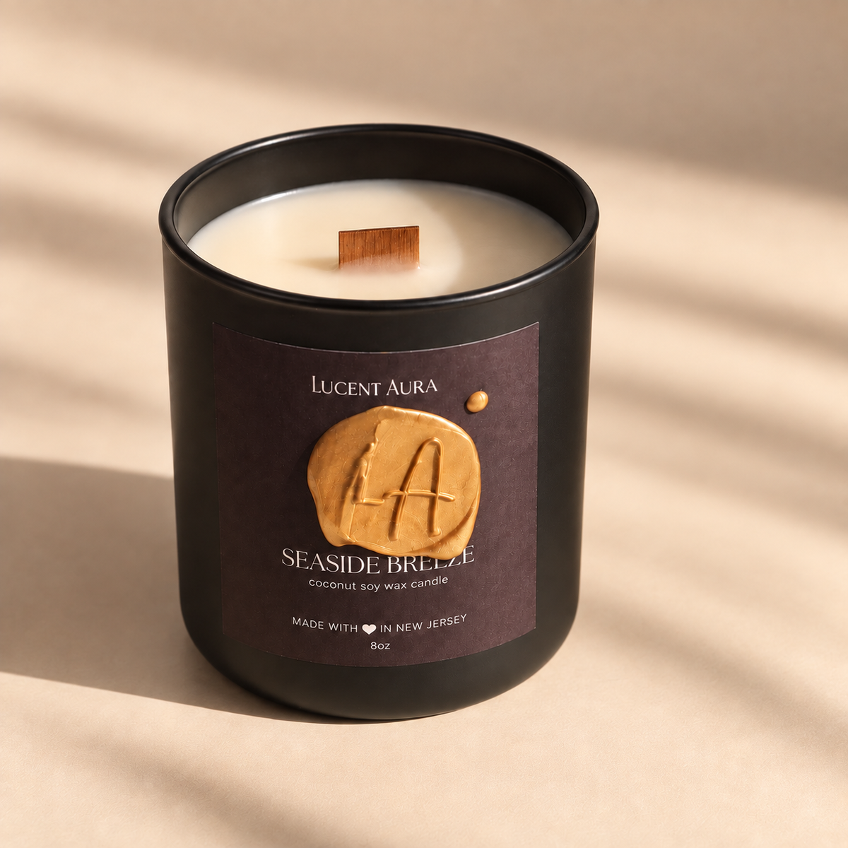 A matte black scented candle jar with cream-colored wax and a small wooden wick, featuring a dark brown label with a gold wax seal and the brand name 'Lucent Aura'. The candle is photographed on a warm beige surface with soft diagonal shadows.
