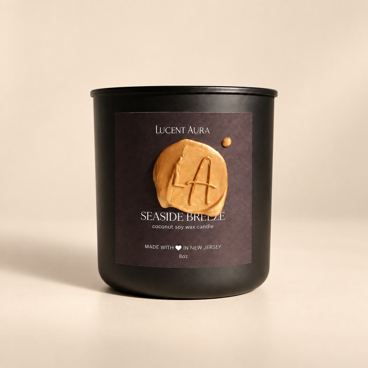 A matte black candle jar with a dark brown label reading 'Lucent Aura' and 'Seaside Breeze', featuring a raised tan wax seal with a letter A, on a warm beige background.