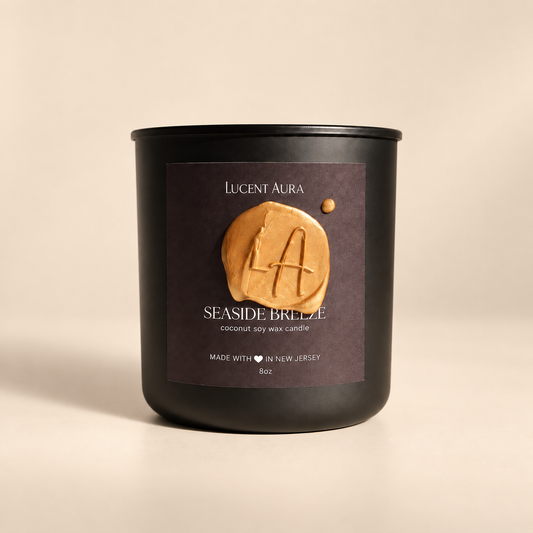 A matte black candle jar with a dark brown label reading 'Lucent Aura' and 'Seaside Breeze', featuring a raised tan wax seal with a letter A, on a warm beige background.