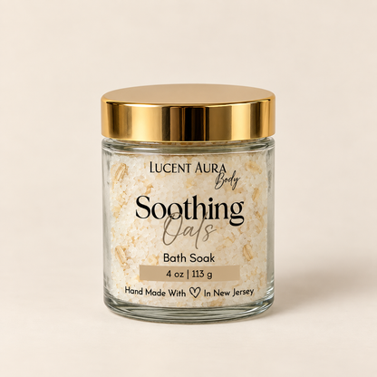 A glass jar of Lucent Aura Body Soothing Oats Bath Soak with a metallic gold lid, set against a solid light beige background.
