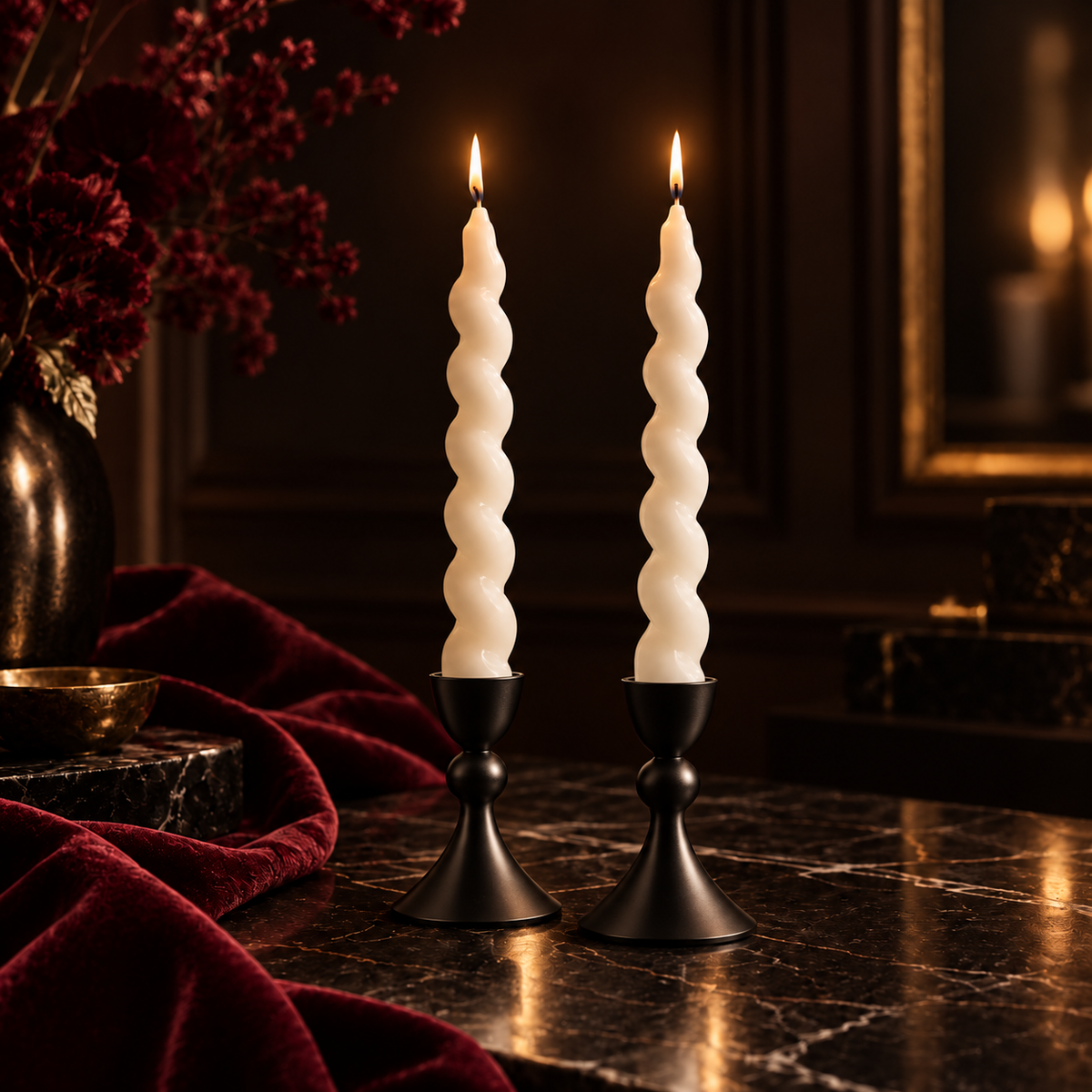 Two lit spiral taper candles in black metal holders on a dark marble table, with deep red velvet fabric and dried flowers in the background.