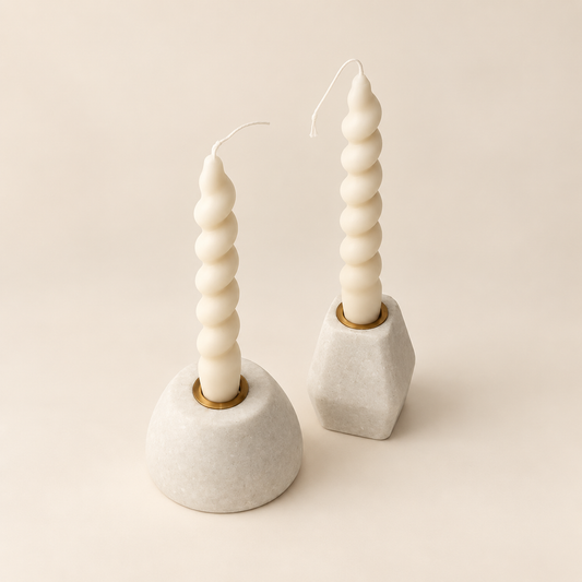 Two sculptural spiral taper candles in matte off-white, displayed in modern geometric holders on a warm beige background.