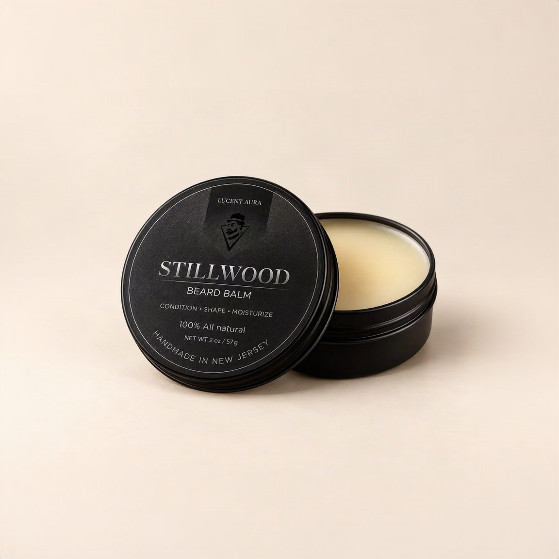 A black round tin of Stillwood Beard Balm by Lucent Aura, with the lid leaning against the open tin showing the pale cream-colored balm inside, set against a solid beige background.