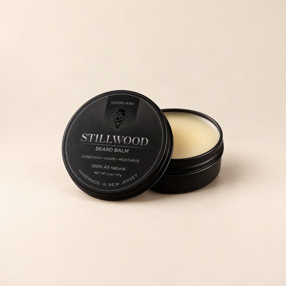 A black round tin of Stillwood Beard Balm by Lucent Aura, with the lid leaning against the open tin showing the pale cream-colored balm inside, set against a solid beige background.