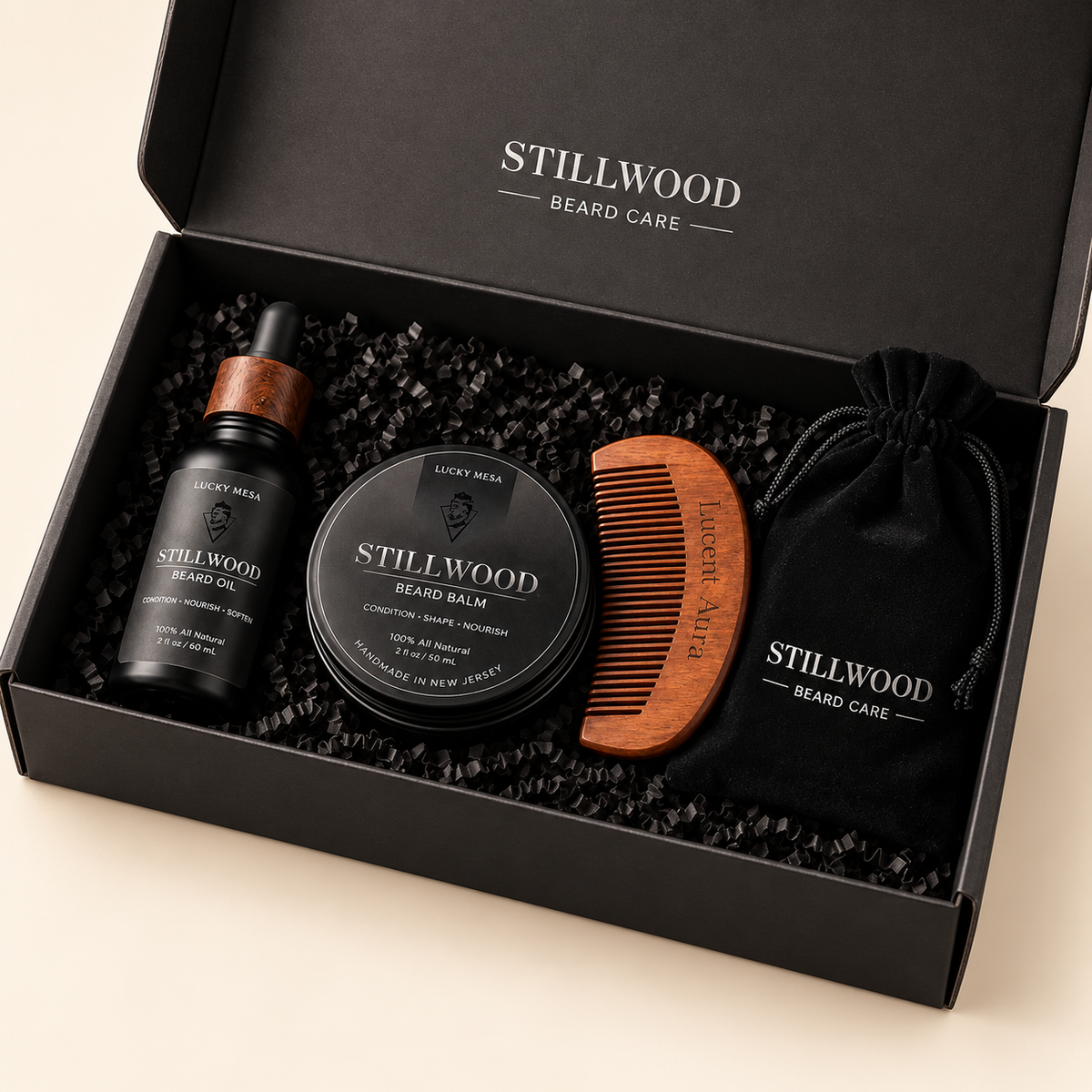 A black gift box from Stillwood Beard Care filled with beard grooming items, including a beard balm tin, a dropper bottle, a wooden comb, and a black drawstring pouch, photographed on a light beige background.