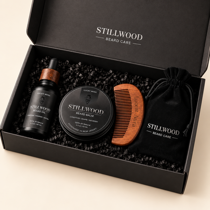 A black gift box from Stillwood Beard Care filled with beard grooming items, including a beard balm tin, a dropper bottle, a wooden comb, and a black drawstring pouch, photographed on a light beige background.