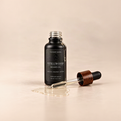 A bottle of Lucent Aura Stillwood Beard Oil with its glass dropper and wooden cap placed beside it on a beige surface.