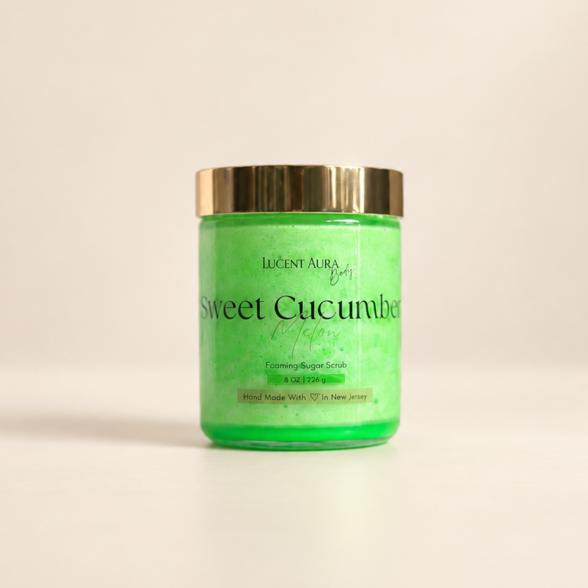 A jar of Lucent Aura Body Sweet Cucumber Melon sugar scrub, featuring a bright green scrub inside, a gold metallic lid, and a clear label with black text, set against a plain off-white background.