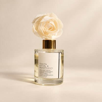 A clear glass reed diffuser bottle with a white label reading 'Vanilla Clouds & Moonflowers' and a large cream fabric flower on a gold cap, set against a warm beige background.