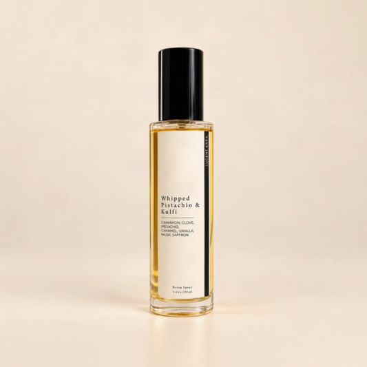 A bottle of Whipped Pistachio & Kulfi room spray, featuring a clear glass bottle filled with golden-yellow liquid, a white label with black text, and a glossy black cap, set against a solid beige background.