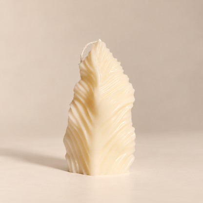 A sculptural leaf-shaped candle in a warm cream or ivory tone, standing upright on a plain beige surface against a matching neutral background.