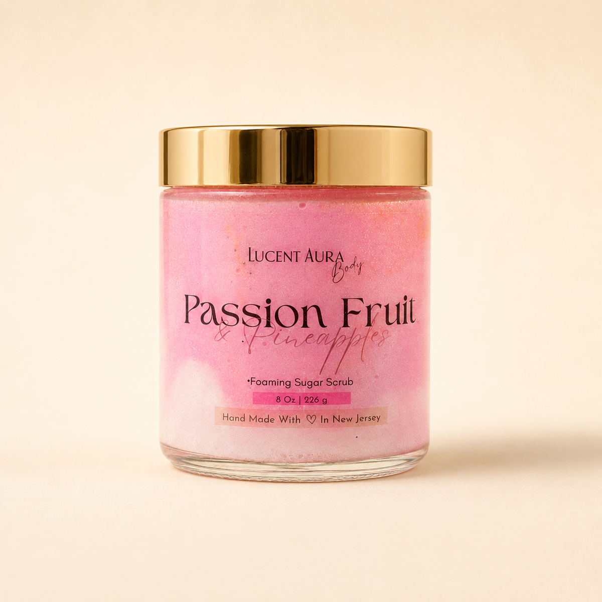 A glass jar of Lucent Aura Body Passion Fruit & Pineapples Foaming Sugar Scrub with a shiny gold lid, set against a solid cream background.