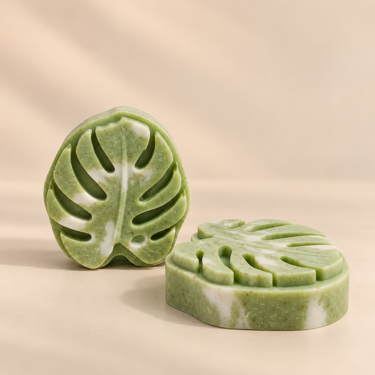 Two green marbled soap bars shaped like monstera leaves, displayed on a warm beige background.