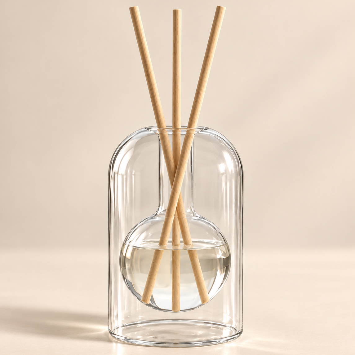 A clear glass reed diffuser with a rounded rectangular body, a spherical water chamber, and four natural wood reeds, set against a warm beige background.