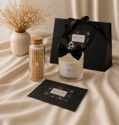 A luxury gift set featuring a clear glass jar candle with a black ribbon bow and silver glitter, a glass jar of wooden toothpicks, and a black gift box and card set, all arranged on a beige satin fabric background.