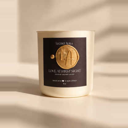 A Lucent Aura soy wax candle in a cream-colored glass jar with a black label featuring a gold wax seal design. The candle is photographed on a warm beige surface with soft, artistic shadows.
