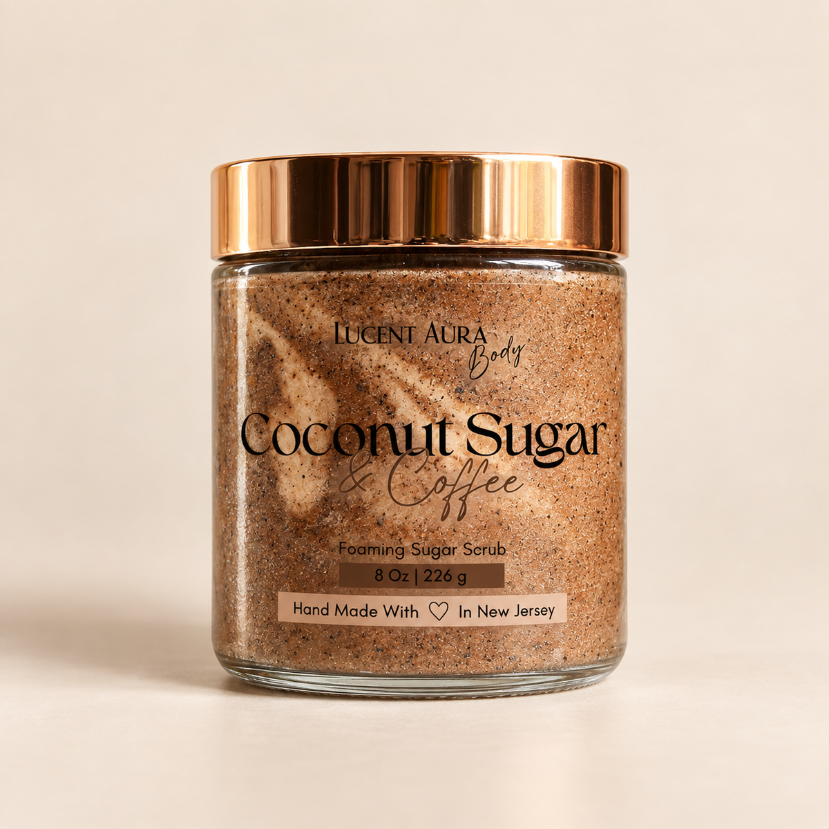 A glass jar of Lucent Aura Body Coconut Sugar Coffee Foaming Sugar Scrub with a metallic rose gold lid, set against a warm beige background.