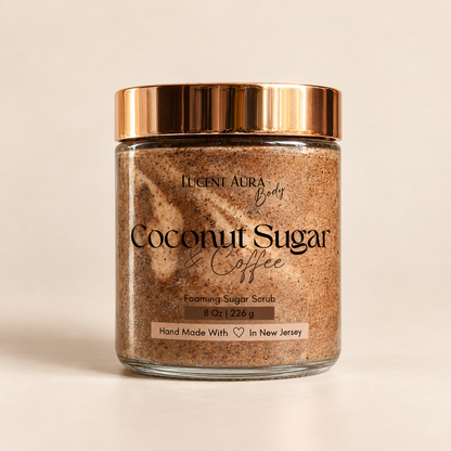 A glass jar of Lucent Aura Body Coconut Sugar Coffee Foaming Sugar Scrub with a metallic rose gold lid, set against a warm beige background.